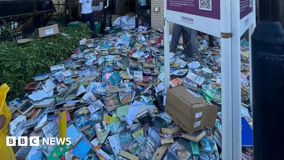 Piles of books cover the pavement outside Broad Green Library in Croydon, many in good condition and some in open boxes. Two people are visible among the stacks, with a black skip to the left and a library sign in the foreground.