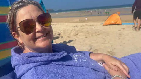 A woman in a blue towelling robe and dark glasses sits on a beach with the sea and a beach shelter behind her.