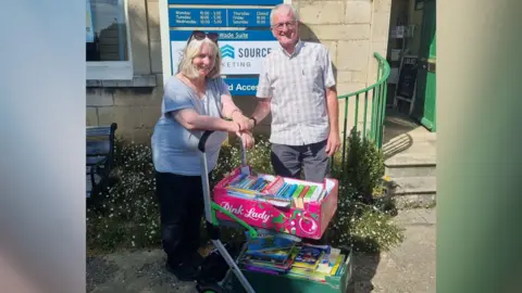 Friends of Woodston Library Volunteers from Deeping Library- a lady wearing a grey T-shirt and Black trousers and a man wearing a checked shirt standing behind a trolley with two crates of books 