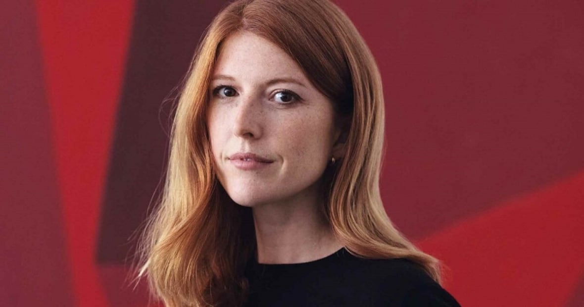 A white woman with red hair looks at the camera against a red background.