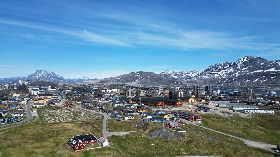 A town located in a valley is shown from a distance at an elevated view, with snow-capped mountains in the background beyond the town.