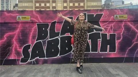 Kirsty Bosley Kirsty Bosley pictured with her arms spread wide and smiling. She is stood against the black and pink Black sabbath wall mural in a cheetah print jumpsuit with chest length hair.