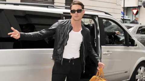 A man wearing a black leather jacket, black sunglasses, a white vest and black joggers, stands beside a silver people carrier with its sliding door open. His right arm is stretched out in a pose and in his left hand he is holding a yellow leather yellow bag. He is standing in a London Street, with other cars and an unreadable street sign in the background.