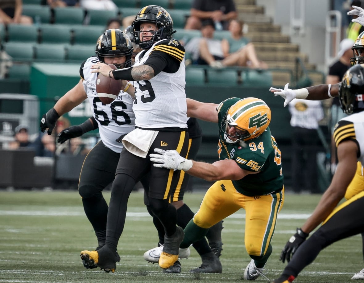 A white football player in a green-and-yellow uniform is falling toward the ground, while trying to wrap his arms around his opponent, wearing a white-and-black uniform. The opposing player has the football cocked in their throwing hand, looking downfield.