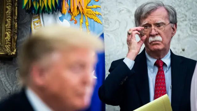 John Bolton (R) adjusts his glasses as he observes Donald Trump (L, blurred in the foreground) from the sidelines during a meeting inside the Oval Office