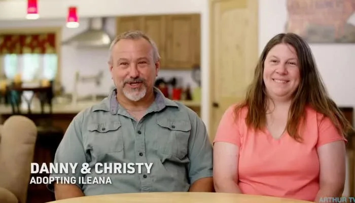 Danny and Christy sit together smiling in a living room setting, casually dressed — text reads "Danny & Christy — Adopting Ileana"