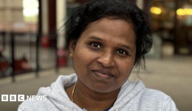Janet is wearing a light grey jumper and has gold earrings and chain.