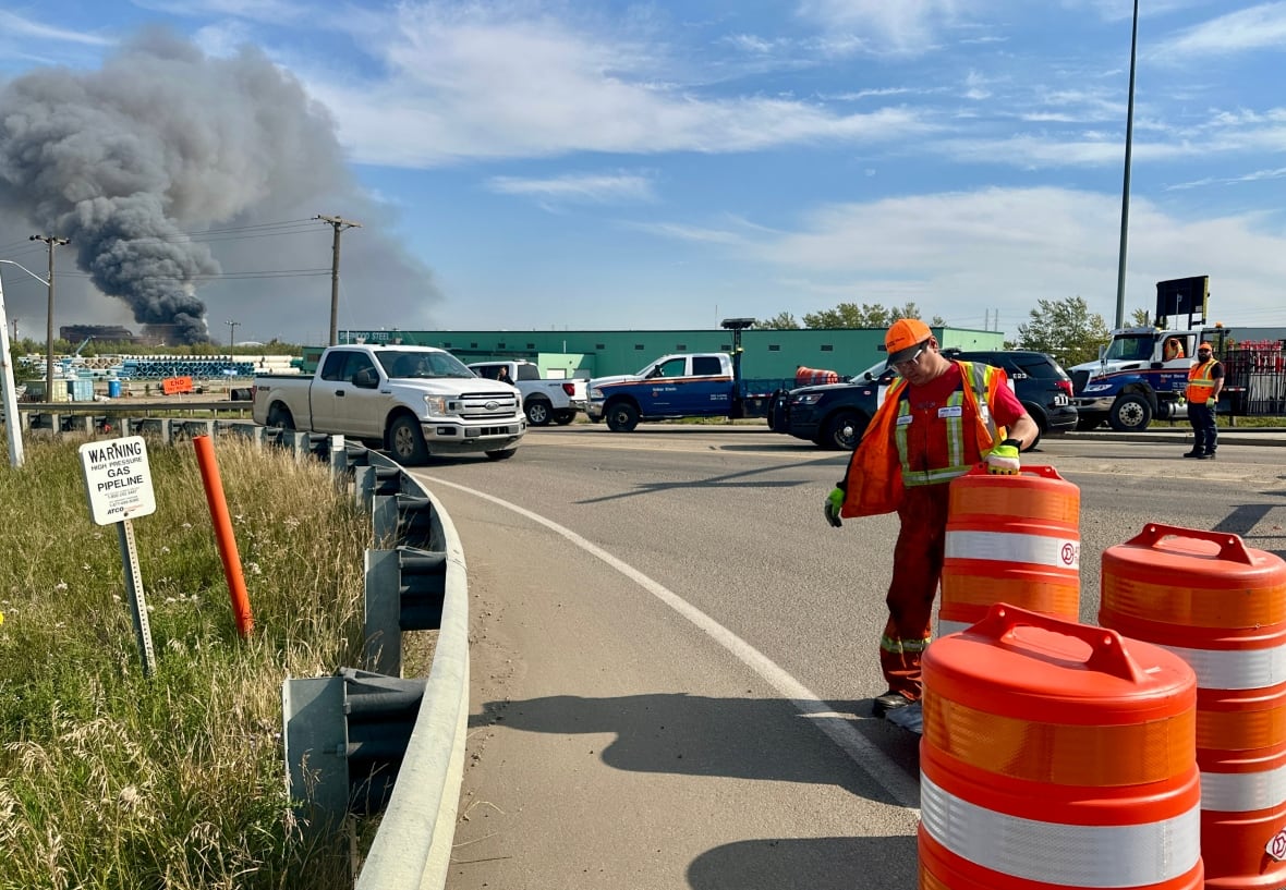 A firefighter moves a pylon on a road. A large plume of smoke can be seen in the background. 