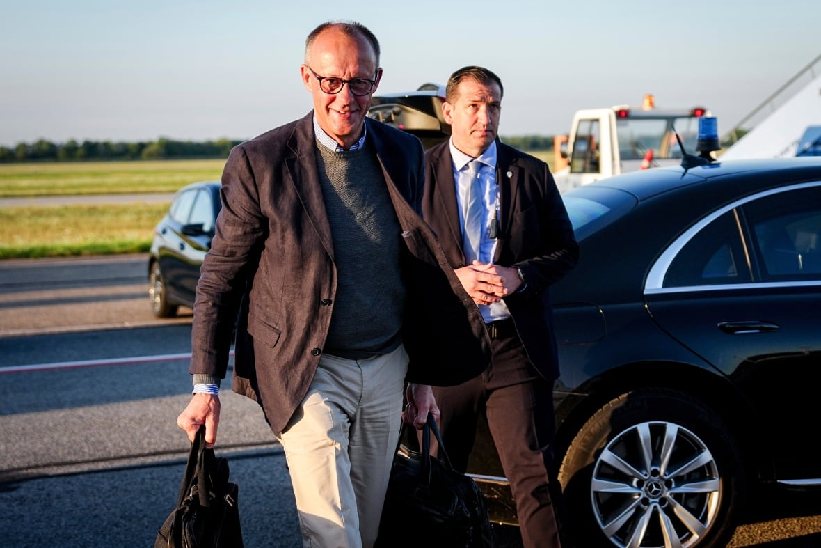 A bespectacled, balding and cleanshaven man is shown walking near a vehicle. Part of an airplane is shown.