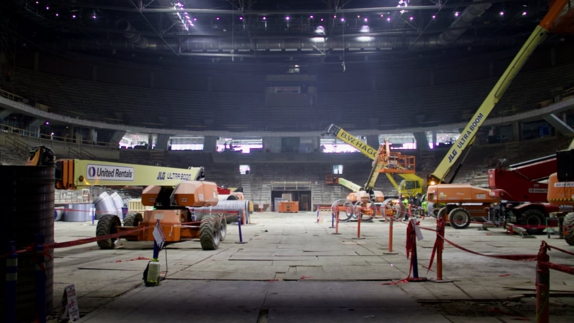 An arena under construction as seen from ground level. There is dust in the air, and cranes parked where the ice surface would be. 