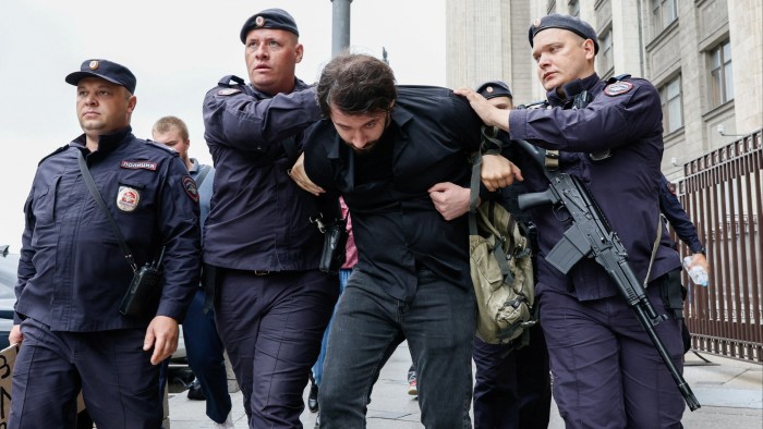 Three policemen in uniform lead away a bearded man in jeans and a black shirt