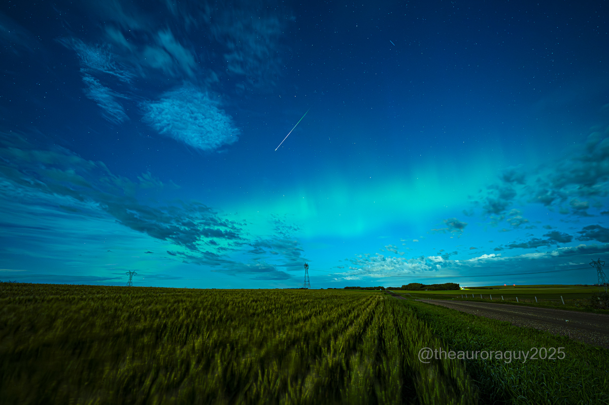 Meteors are visible streaking through a dark blue sky wreathed in a green aurora above a rural landscape.