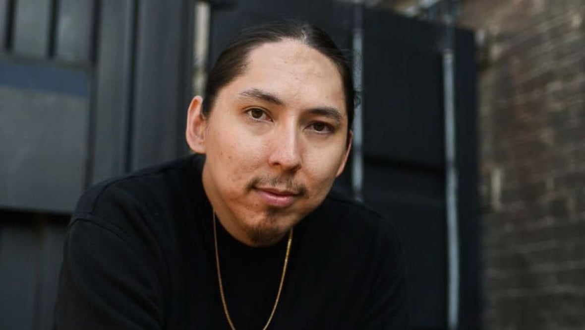 A headshot of of a man with a goatee and his hair tied back wearing a black shirt and a gold chain.
