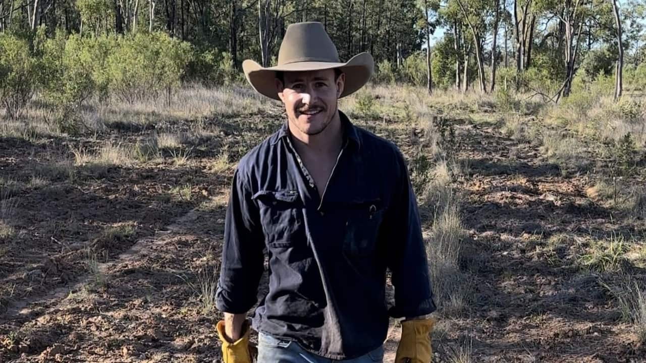 A young man wearing a dark blue long-sleeved shirt, Akubra hat and gloves walks in a bushland setting