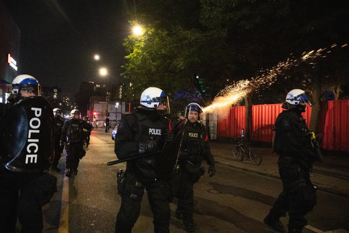 A group of police officers in riot gear walking on a street, a line of sparks over their heads