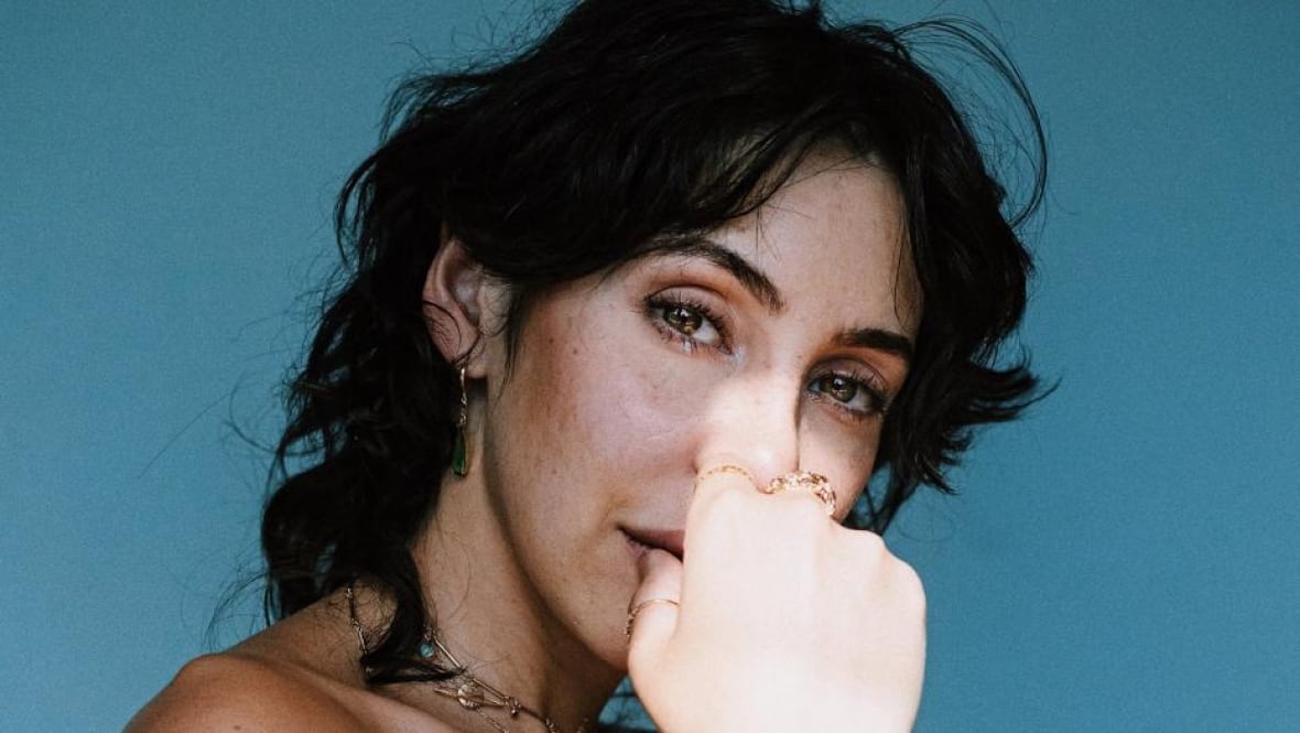 A headshot of a person with black curly hair with their hand in front of their mouth against a blue background.