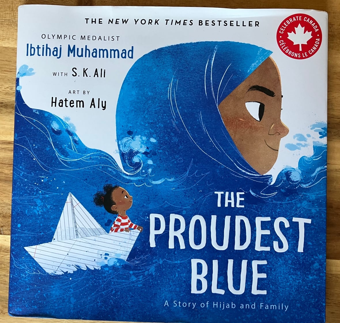 A book called 'The Proudest Blue' with a small girl in a paper boat, sailing in waves which create a hijab that her older sister is wearing. 