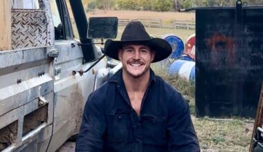 a young man in a navy button-up shirt and an Akubra hat smiles at the camera while sitting in a paddock on a farm next to a ute