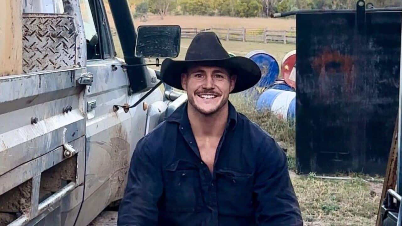a young man in a navy button-up shirt and an Akubra hat smiles at the camera while sitting in a paddock on a farm next to a ute
