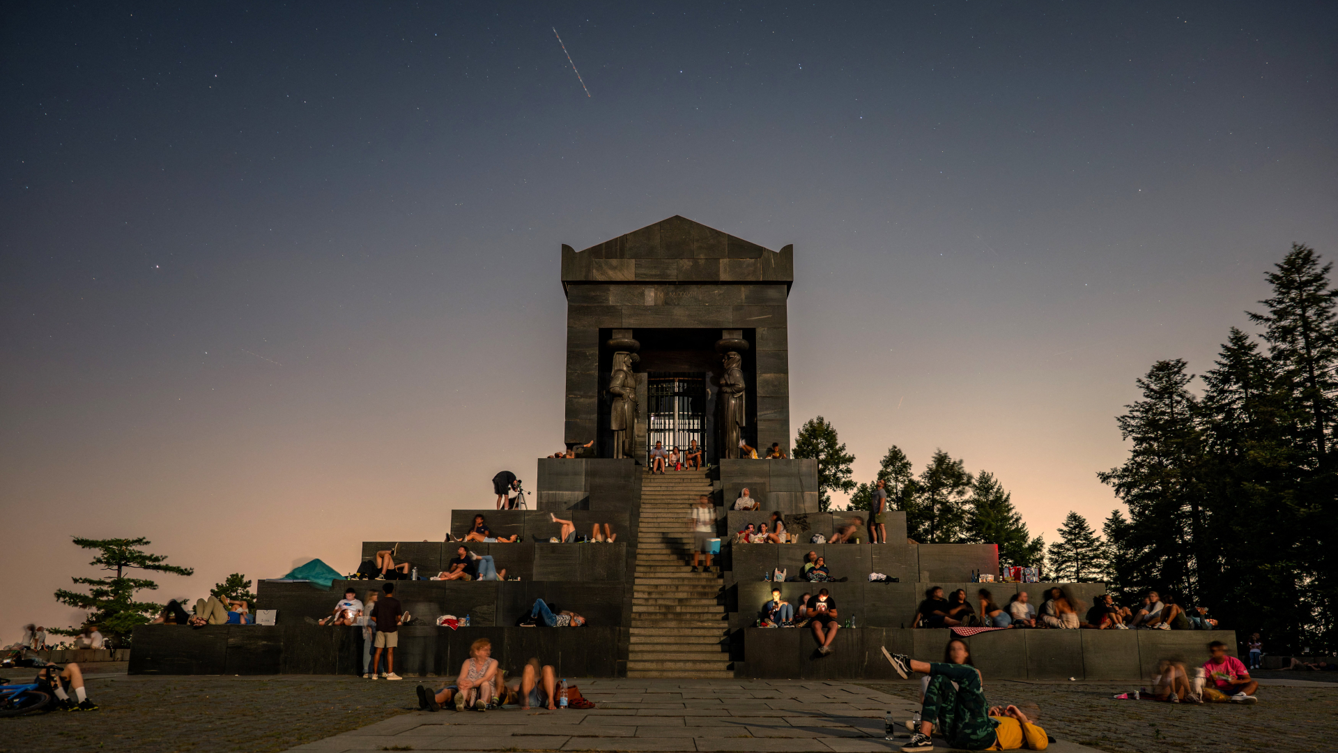 A meteor streaks through the sky above a stepped temple where a crowd of stargazers has gathered.