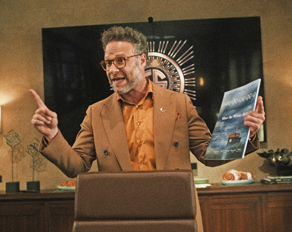 Man in a tan suit and orange shirt holds a book and points while standing behind a podium.