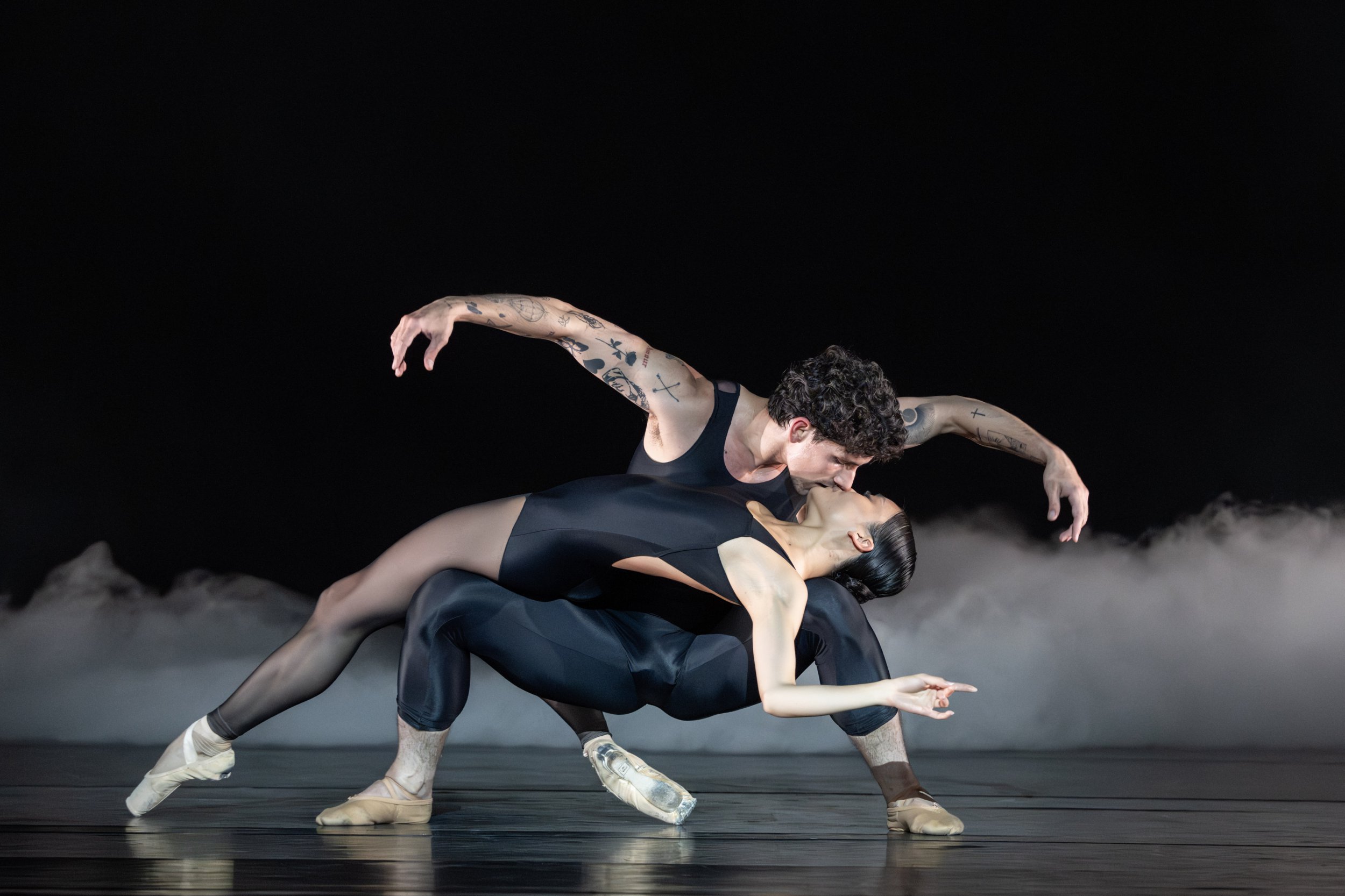 Two ballet dancers in black leotards kissing during a performance.