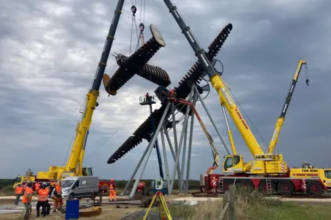 Two sections of a large rusted steel structure, in the shape of a World War Two bomber aircraft, are lifted on to a steel support by two large yellow cranes as workers in orange and yellow hi-vis jackets watch from the ground. The sculpture is seen from behind and the afternoon sky is darkening.