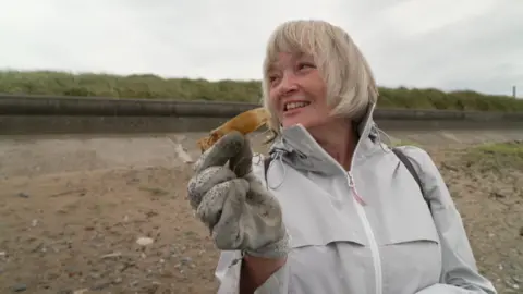 Heather Neal is wearing a grey jacket and gloves and is holding an old shark egg case in her right hand. She is standing on the sand at Morecambe Bay.