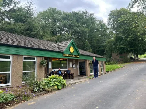 A man stands outside Melrose Golf Club club house with a few people standing outside. It is a low, single-story building with green wood panels and the yellow name on black of the golf club.