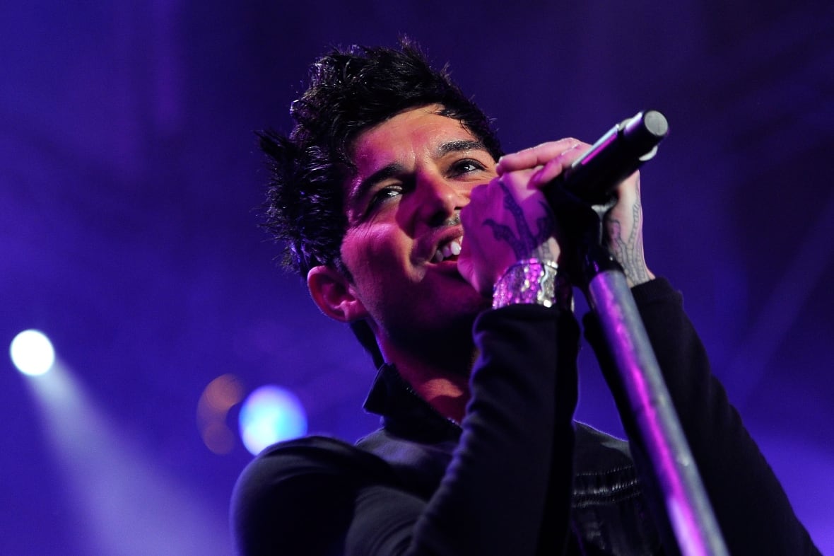 A man wearing black sings on stage while surrounded by purple lighting in the background.