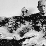 American abstract expressionist painter Jackson Pollock (1912 - 1956) holds a cigarette above as he stands behind a painting in his studio at 'The Springs,' East Hampton, New York, August 23, 1953. (Photo Tony Vaccaro/Getty Images)