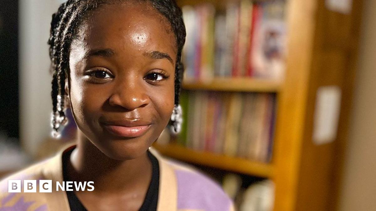 Hephzibah Akinwale who has dark hair in short braids pulled back from her face and is starting to smile. She is wearing a pink and yellow cardigan over a black top. Behind her is a bookshelf full of books.