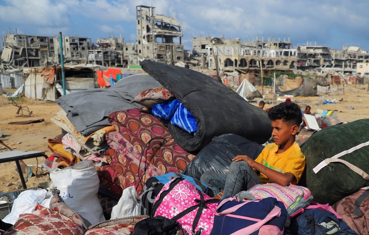 A boy sits on top of a pile of belongings in front of destroyed buildings.
