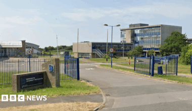 Google Street View of Budmouth Academy in Weymouth. Blue metal gates open onto a drive surrounded by buildings. The largest is a late 20th-century four-storey block covered in glass and blue panels and with a flat roof.