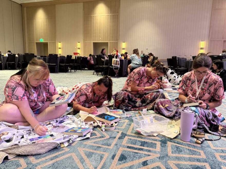 A group of friends bejewel the covers of their favorite romance novels while clad in book-themed pajamas during the Romantically Yours Convention’s pajama party event. (Hafsa Quraishi/Here & Now)