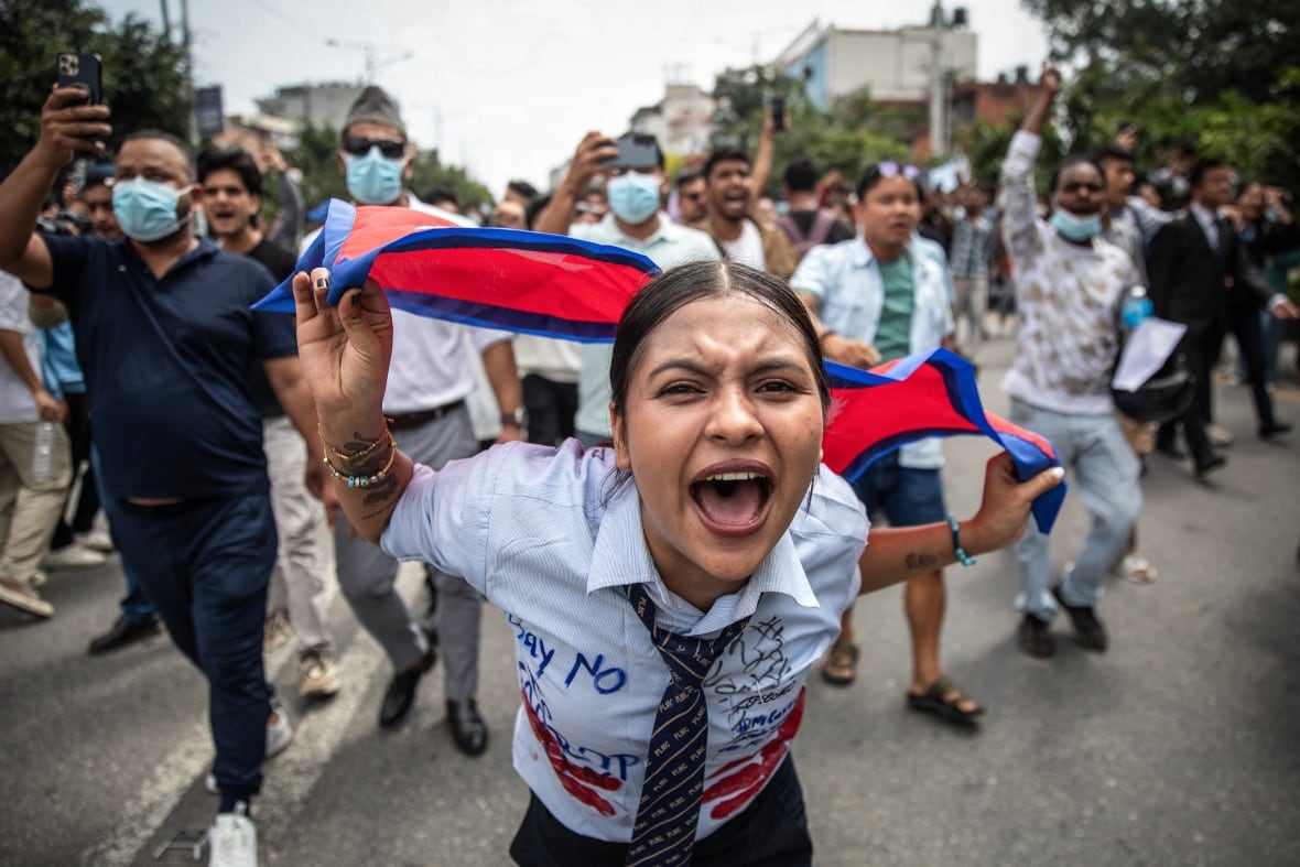A young woman close to the camera wearing a shirt and tie appears to yell while carrying a flag, as others follow behind her in an apparent street post.