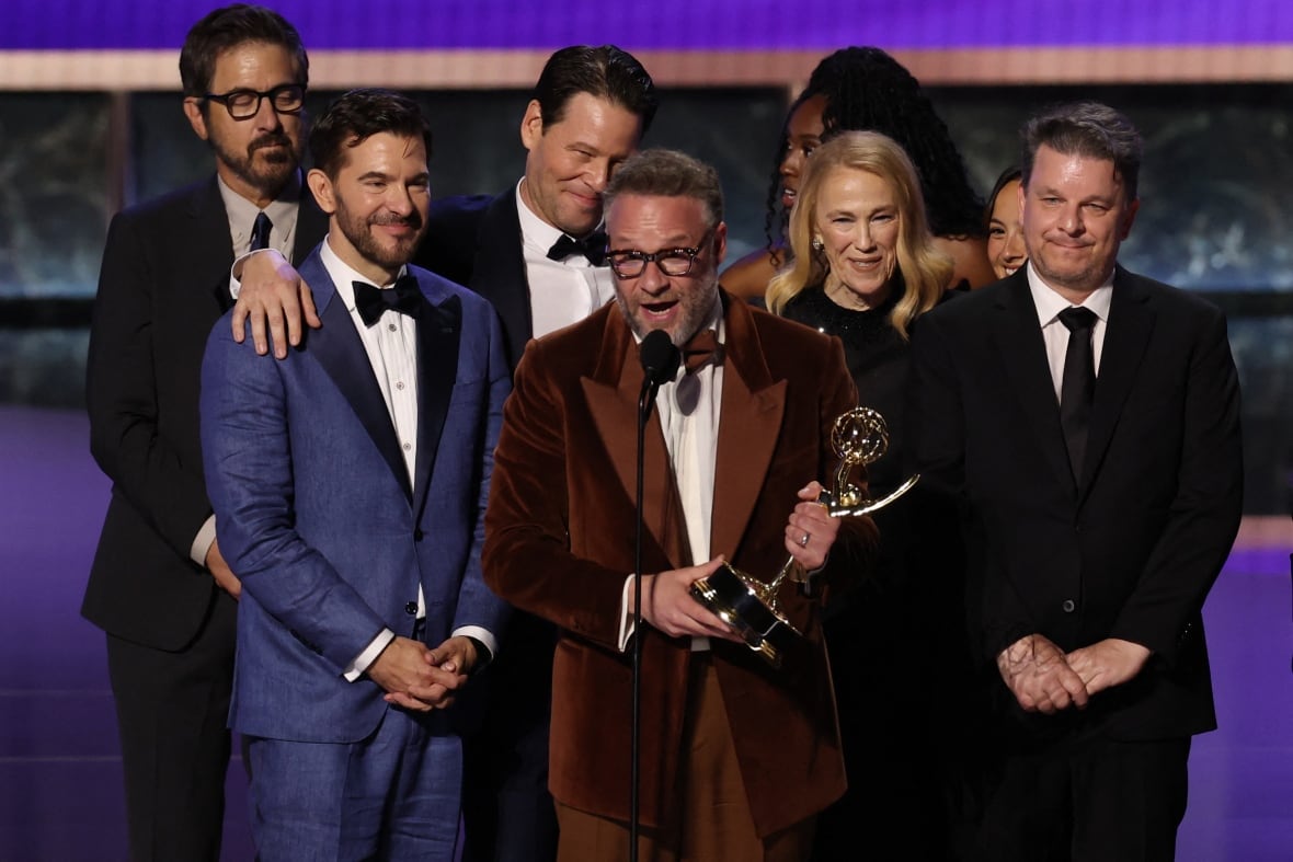 Bearded man holding an Emmy award with others behind him on stage making an acceptance speech.