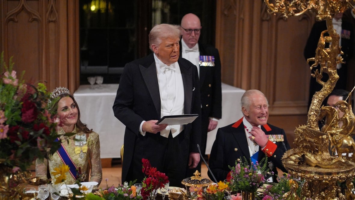 An elderly man standing up while two people, a woman in gown, sash and tiara and an elderly man grimacing and adjusting his neck collar. They're at an elaborate long dining table during a formal banquet.