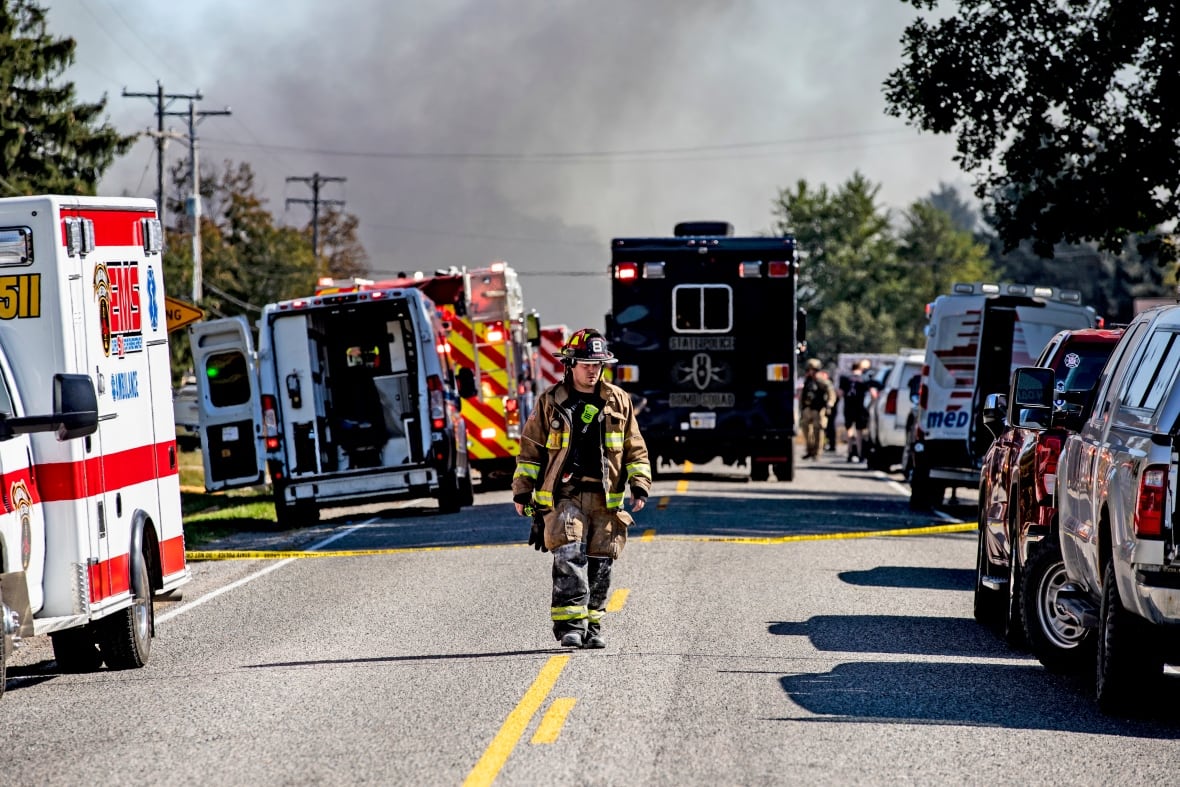 A firefighter walks down the street. He's surrounded by emergency service vehicles, and black smoke rises in the background.