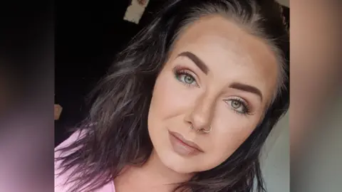 Sammi Hill A selfie of Sammi Hill, a 33-year-old woman with dark brown hair. She is wearing a full face of make up and a pink t-shirt. She is subtly smiling. 