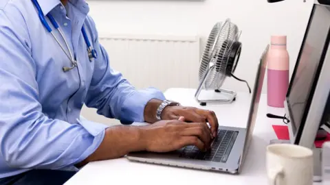 Getty Images Doctor in a blue shirt with a stethescope round his neck looking at a laptop at a desk with a monitor. You can only see the body and not the head