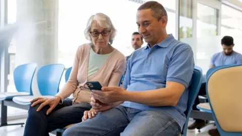 Getty Images A waiting area with several people seated on blue and beige chairs. In the foreground, a man and a woman are sitting together and looking at a smartphone.