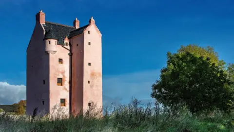 Landmark A view of Fairburn Tower, a building which has been rendered in a pink-colour. The tower has tiny windows over five levels , with a circular turret and grass grows wild around it as it stands against the blue sky.
