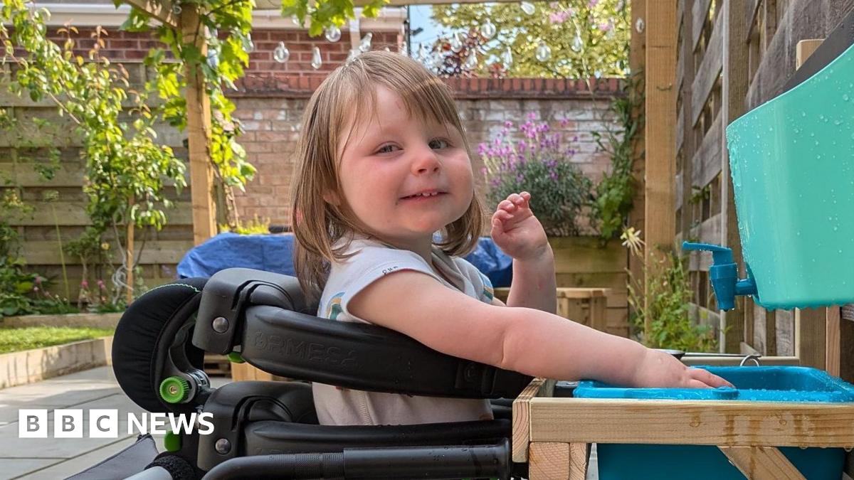 A three-year-old girl in a wheelchair playing with water. She is smiling at the camera. She has long brown hair and a fringe. She is in a garden on a sunny day.