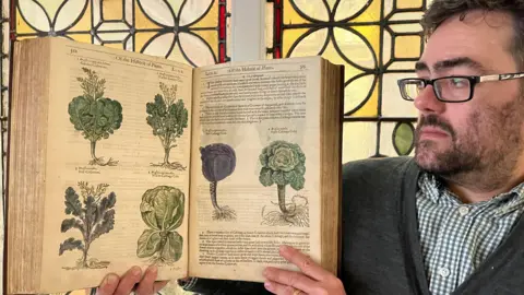Leeds City Council A man holding up an open book the camera. The book is old and features illustrations of plants. The man is on the right of the picture. He has glasses and is wearing a blue and white checked shirt and a grey cardigan.