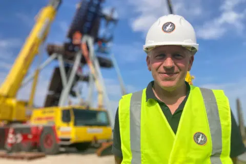 A man wearing a yellow hi-vis jacked and white hard hat smiles as he stands in front of a large yellow crane and steel sculpture, which are out of focus in the background beneath a blue sky.