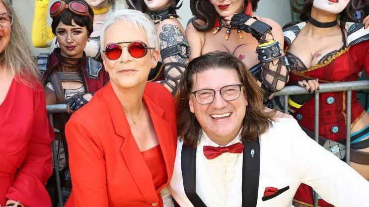 A man in a white tuxedo and red bow tie smiles widely next to a woman in a brick red suit and matching sunglasses. Behind them, a row of female cosplayers dressed in futuristic steampunk outfits and face paint inspired by the Borderlands video games pull sultry poses.