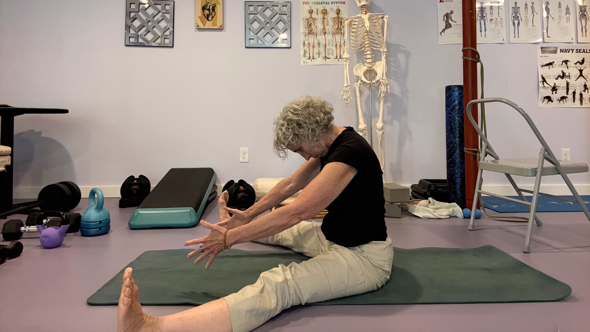woman in light trousers and dark top sat on the floor legs outstretched wide on a green mat in a studio setting.