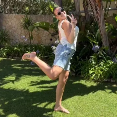 Grace Parkin @forevergraceful21 A woman in a sleeveless white top, denim shorts and sandals poses in a garden for a photo.