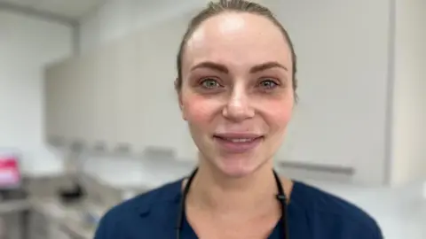 May is stood in a dentist's surgery wearing a navy blue top with her glasses' chain visible around her neck. Her hair is tied back off her face and she is smiling at the camera.